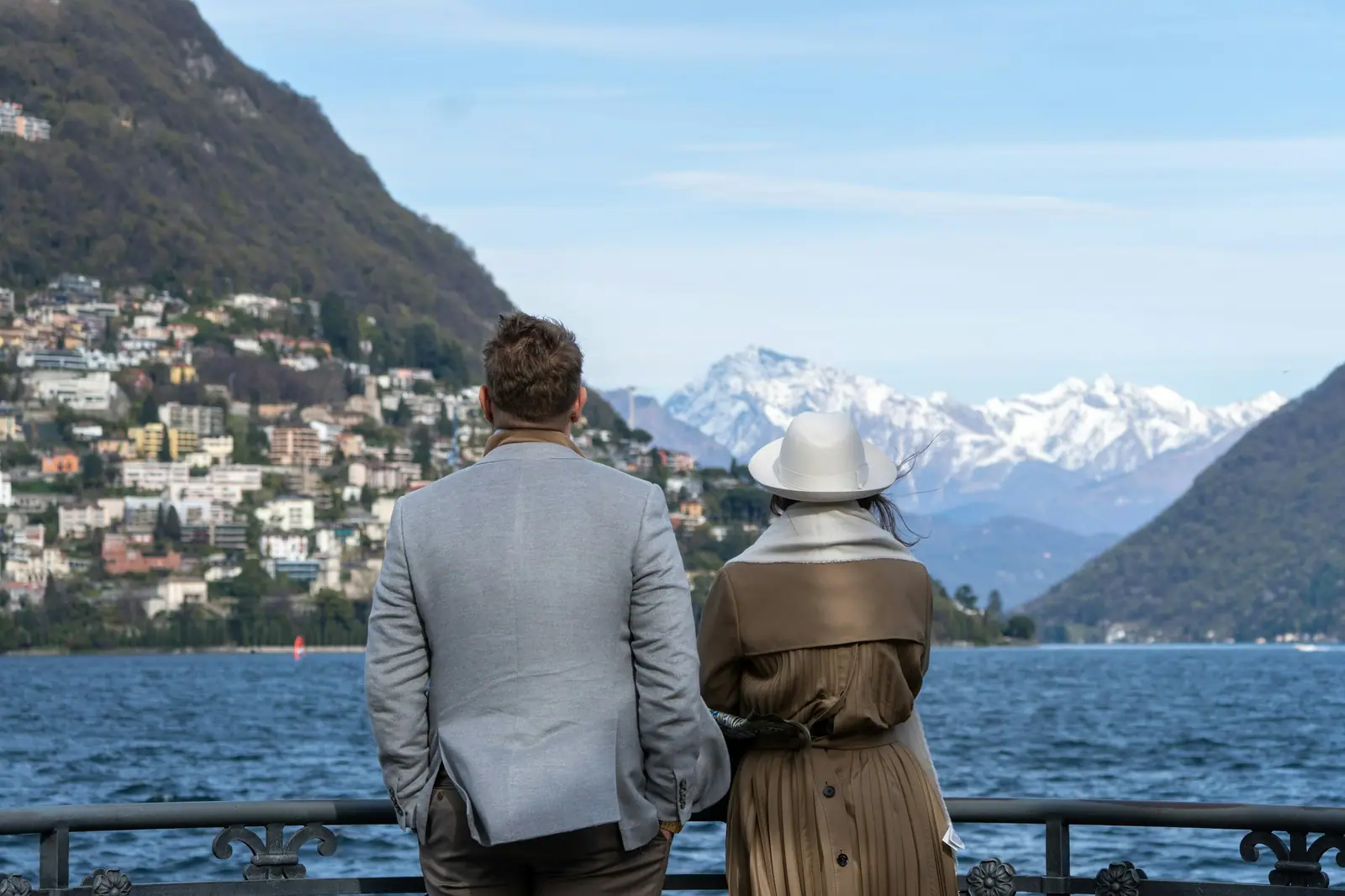 Ein Paar steht an der Seepromenade von Lugano und blickt über den See auf verschneite Berggipfel